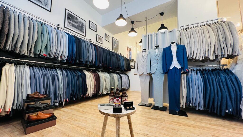 Hochzeitshaus Schubert – Brautmode & Hochzeitsanzüge 9 Herrenabteilung im Hochzeitshaus Schubert mit Hochzeitsanzügen an Kleiderstangen, drei Anzügen an Puppen und Schuhen